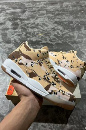 A pair of Jordan 'Desert Camo' lifestyle sneakers, displaying their camouflage pattern, white midsoles with visible Air units, and gum outsoles, resting on a beige shoe box.