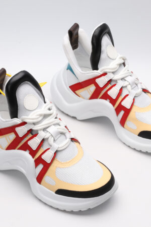 A pair of white chunky athletic sneakers with vibrant orange and light grey overlays, white laces, black accents on the toe and heel, and a large, sculpted white sole, displayed against a light grey background with one shoe showing the sole.