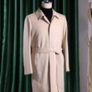 A high-quality men's sand-colored overcoat with a self-tie belt, notched collar, and hidden buttons, displayed on a mannequin against green curtains.