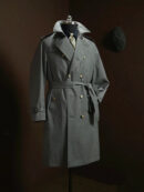A slate grey double-breasted men’s trench coat with a waist belt, wide lapels, and tortoise-shell buttons, displayed on a mannequin against a dark brown background.