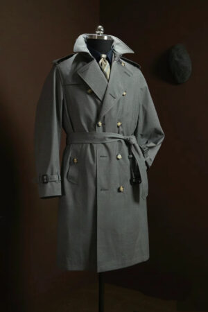 A slate grey double-breasted men’s trench coat with a waist belt, wide lapels, and tortoise-shell buttons, displayed on a mannequin against a dark brown background.