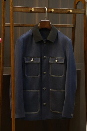 A dark indigo denim safari jacket with a black contrast collar and four front pockets hanging on a wooden rack.