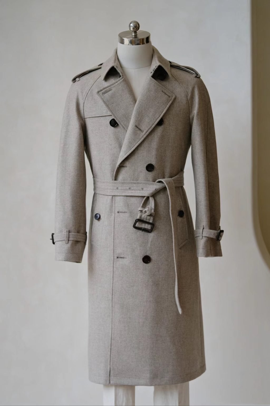 A light oatmeal-colored wool-blend trench coat for men, featuring a double-breasted front and belted waist on a mannequin.