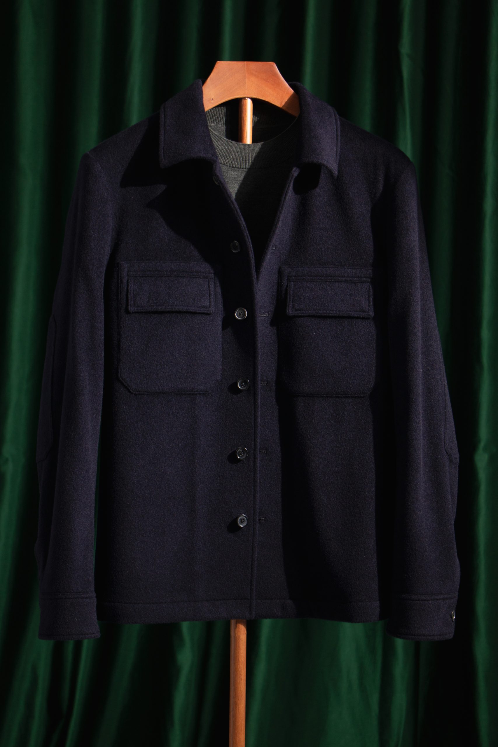 A navy blue wool safari overshirt hanging against a green velvet curtain, featuring two large chest pockets and a simple collar.