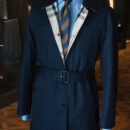 A navy blue men's trench coat with an adjustable waist belt and a visible reversible check pattern on the interior lapels, layered over a blue shirt and striped tie.