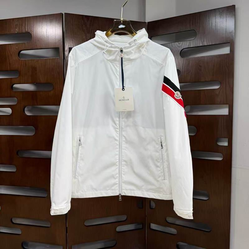 White Moncler Grimpeurs windbreaker jacket with a hood, front zipper, and red and black striped band on the left sleeve hanging on a gold hanger.