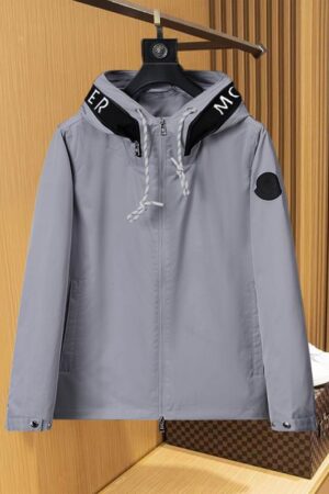 A front view of a grey men's windbreaker with a black-rimmed hood featuring white lettering, hanging against a wooden slat background.