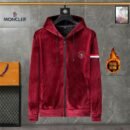 A front view of a burgundy velvet zip-up hoodie with a black zipper, thick drawstrings, a fleece-lined hood, and a white stripe detail on the sleeve, hanging in a modern room setting.