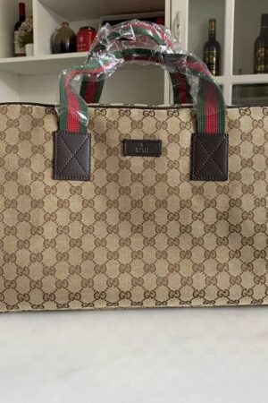 A Gucci Savoy duffle bag with beige GG Supreme canvas, brown leather trim, and green and red striped handles sitting on a white marble surface.