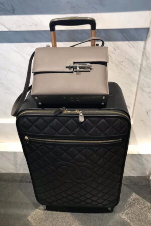 A luxury black quilted leather suitcase with a gold telescopic handle and the iconic interlocking logo design standing against a marble wall.
