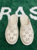 A pair of cream-colored rubber slip-on mules with perforated interlocking logos resting on a green textured rug.