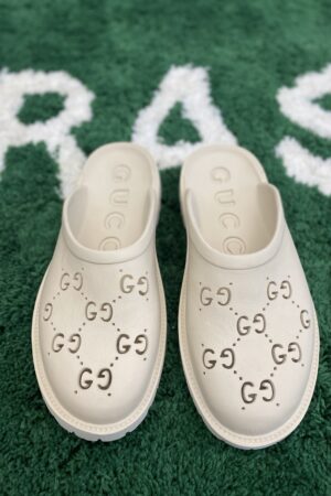 A pair of cream-colored rubber slip-on mules with perforated interlocking logos resting on a green textured rug.