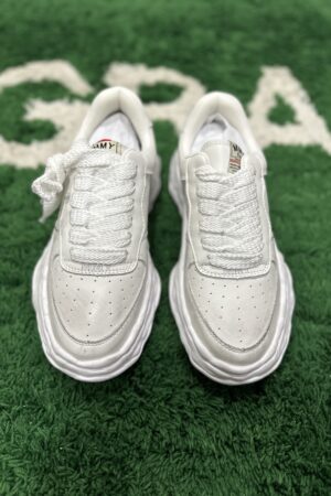 Maison Mihara Yasuhiro Wayne low-top sneakers in all-white leather featuring thick distorted soles, oversized laces, and perforated toe boxes on a green textured background.