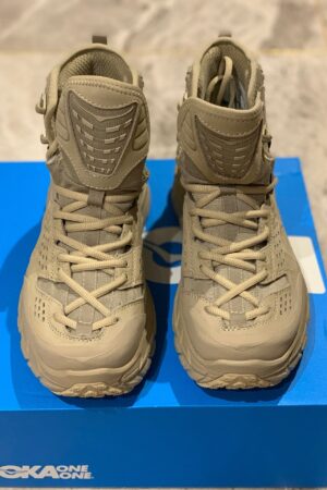 An overhead view of tan Hoka One One x Engineered Garments Tor Ultra Hi boots featuring thick tonal midsoles, high-top ankle support, and a technical lacing system, resting on a gray marble surface.