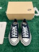 A pair of black Maison Mihara Yasuhiro Peterson low-top sneakers with chunky, warped white midsoles and white laces, displayed on a green textured surface with a tan shoebox in the background.