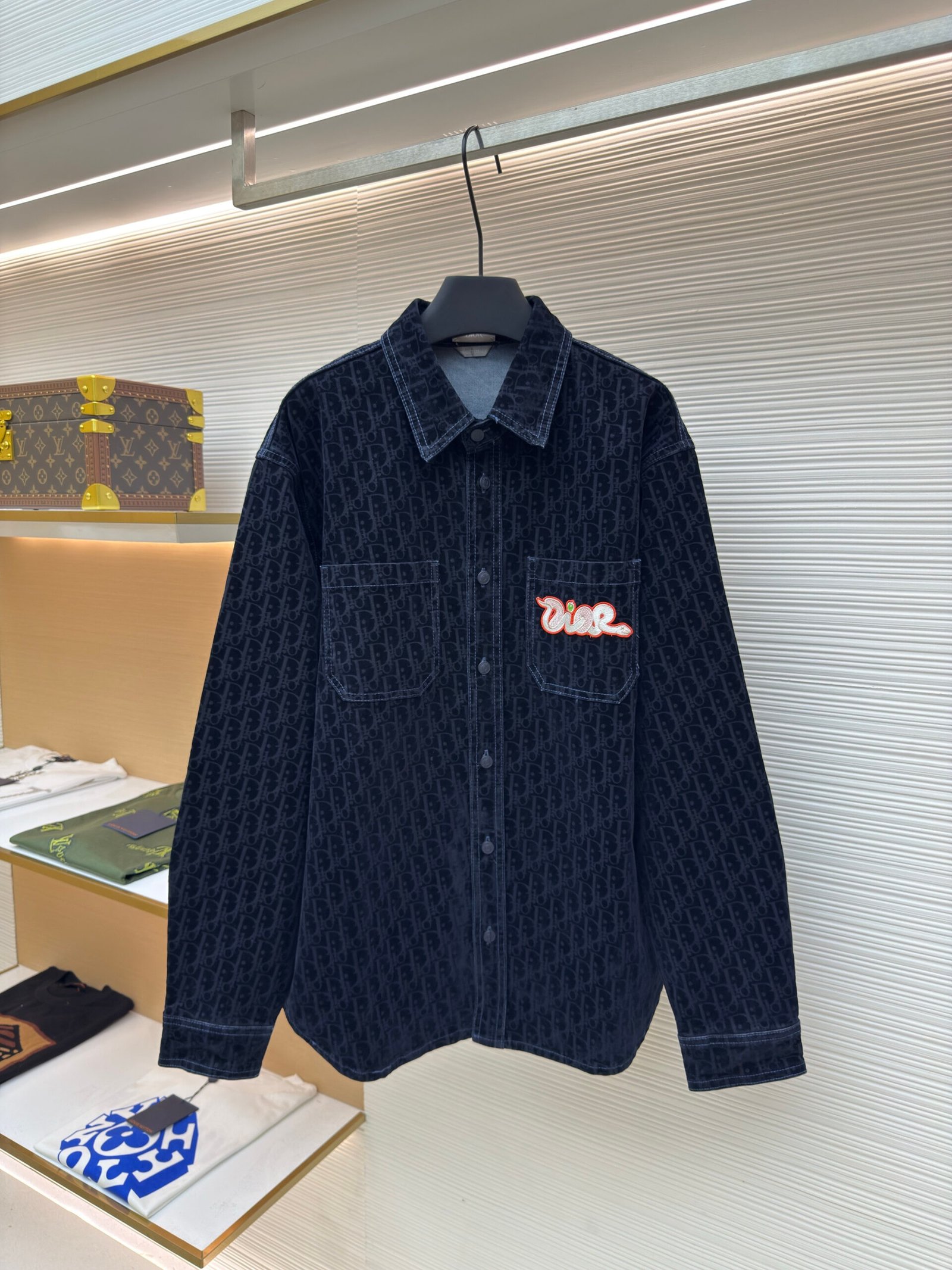 A dark navy denim button-down overshirt featuring an all-over Dior Oblique monogram pattern and a red script "Dior" patch on the chest.