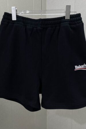 Black Balenciaga fleece shorts hanging on a metal rack, featuring a white embroidered 3B sports icon logo on the left thigh and an elastic drawstring waistband.