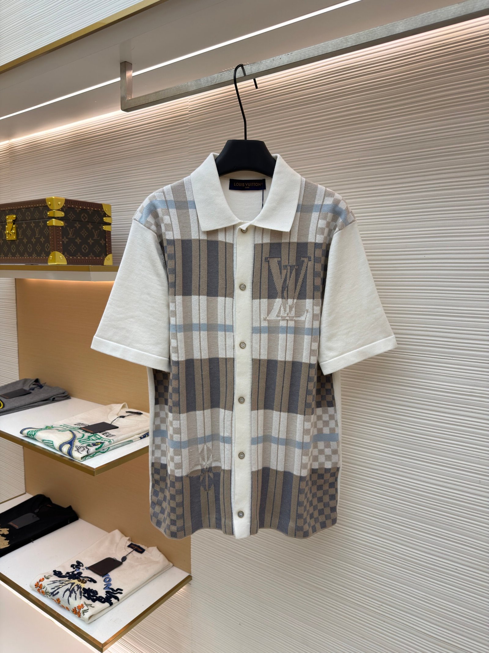 A premium men's checkered knit polo shirt in beige, grey, and cream featuring large "LV" chest embroidery hanging on a display rack.