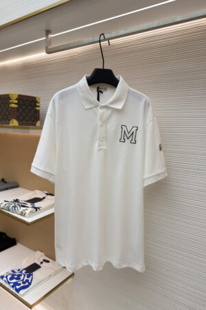 A crisp white men's pique polo shirt featuring a black-outlined varsity "M" on the chest and a designer sleeve patch hanging on a display rack.