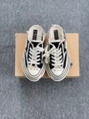 A pair of black and white Maison Mihara Yasuhiro Baker canvas mules featuring a thick, warped white midsole, frayed edges, and oversized laces, displayed next to a tan shoebox.