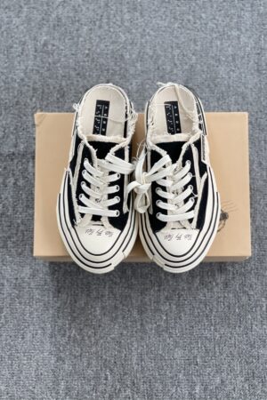 A pair of black and white Maison Mihara Yasuhiro Baker canvas mules featuring a thick, warped white midsole, frayed edges, and oversized laces, displayed next to a tan shoebox.