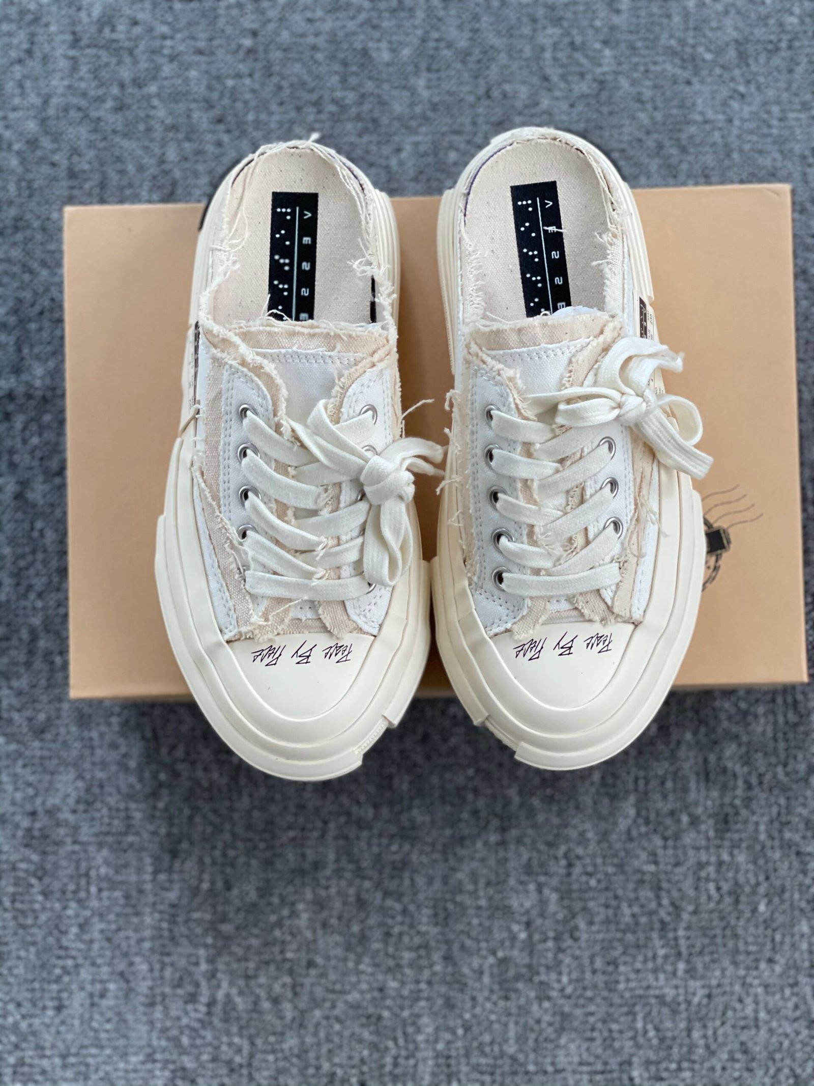 op-down view of white and cream Maison Mihara Yasuhiro Baker canvas mules with chunky warped soles and oversized laces, resting on a tan cardboard box.