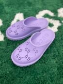 A top-down view of vibrant purple rubber slip-on mules featuring perforated interlocking logos, resting on a green textured rug.