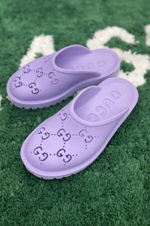 A top-down view of vibrant purple rubber slip-on mules featuring perforated interlocking logos, resting on a green textured rug.