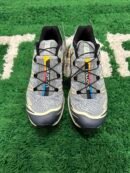 A pair of grey and charcoal Salomon XT-6 trail sneakers with thin black laces, dark geometric overlays, and a vertical yellow, red, and blue stripe on the tongue, displayed on a green textured surface.