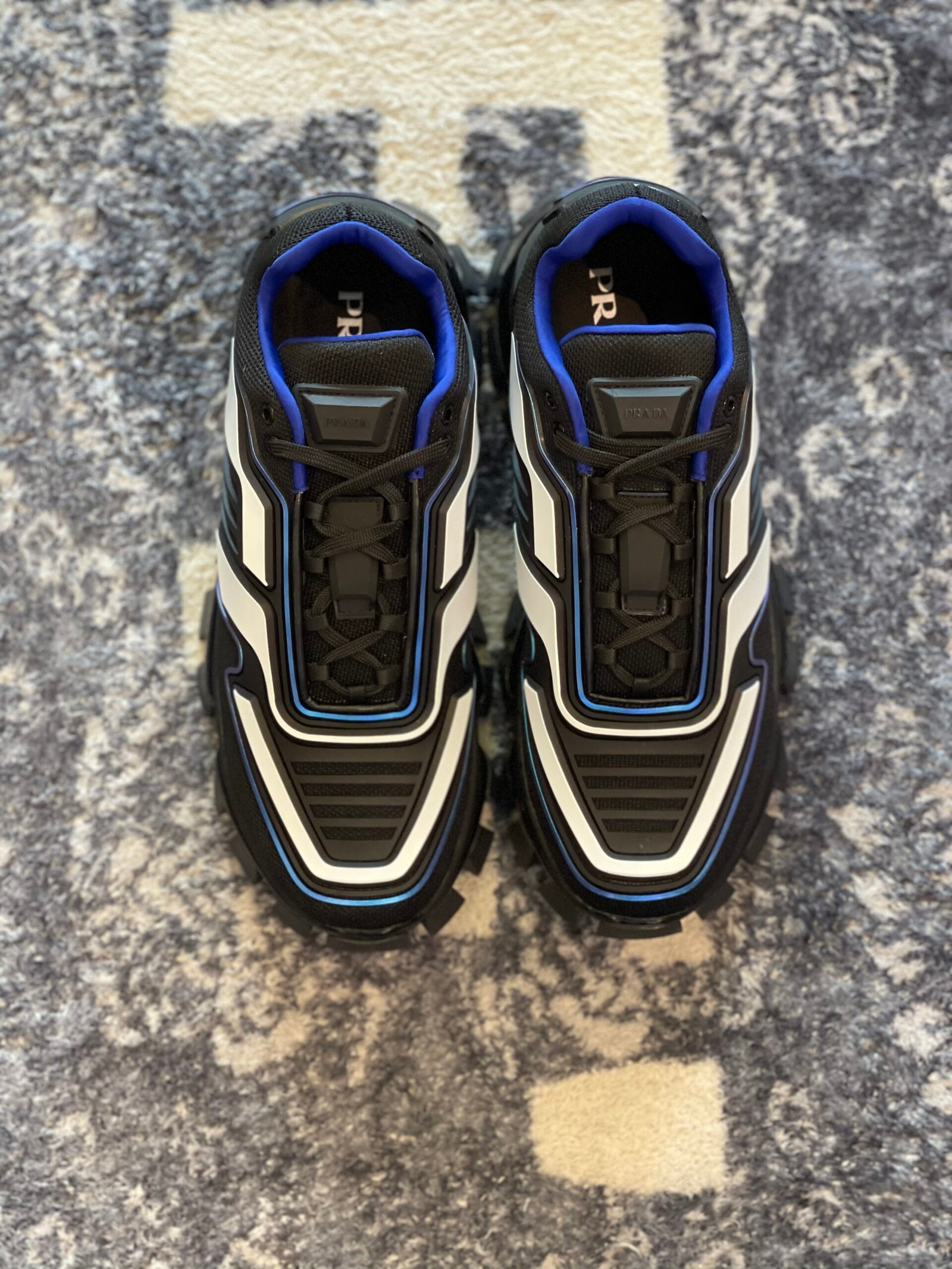 A pair of Prada Cloudbust Thunder sneakers in black with white panels and blue trim on a textured rug.