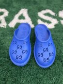 A top-down view of vibrant blue rubber slip-on mules featuring perforated interlocking logos, resting on a green textured rug.