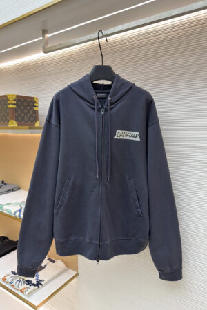 A dark zip-up hoodie featuring a white tape-style Balenciaga logo on the left chest.