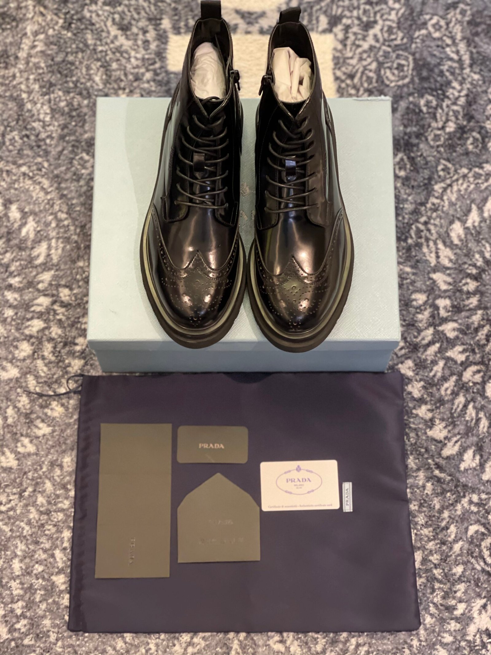 Top view of black Prada Spazzolato leather brogue combat boots displayed on a light blue box with accessories.