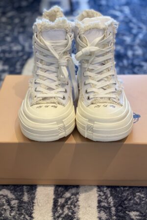 Front view of white Mihara Yasuhiro high-top sneakers with frayed canvas edges and chunky distorted soles on a tan box.