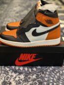 A high-top orange, black, and white leather sneaker resting on a black box with a red logo.