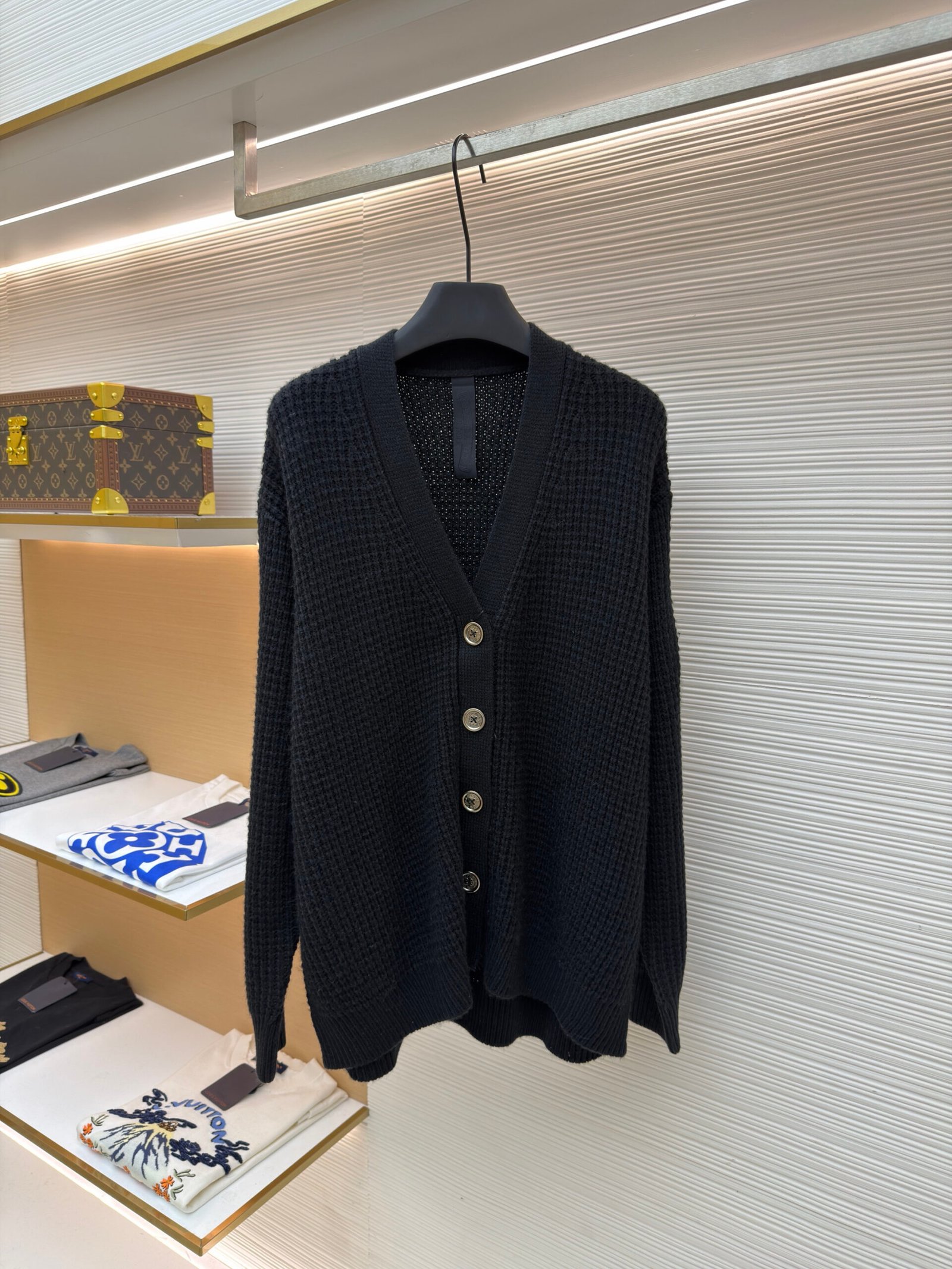 Black Louis Vuitton waffle knit V-neck cardigan hanging on a clothing rack with silver-toned engraved buttons and a textured weave.