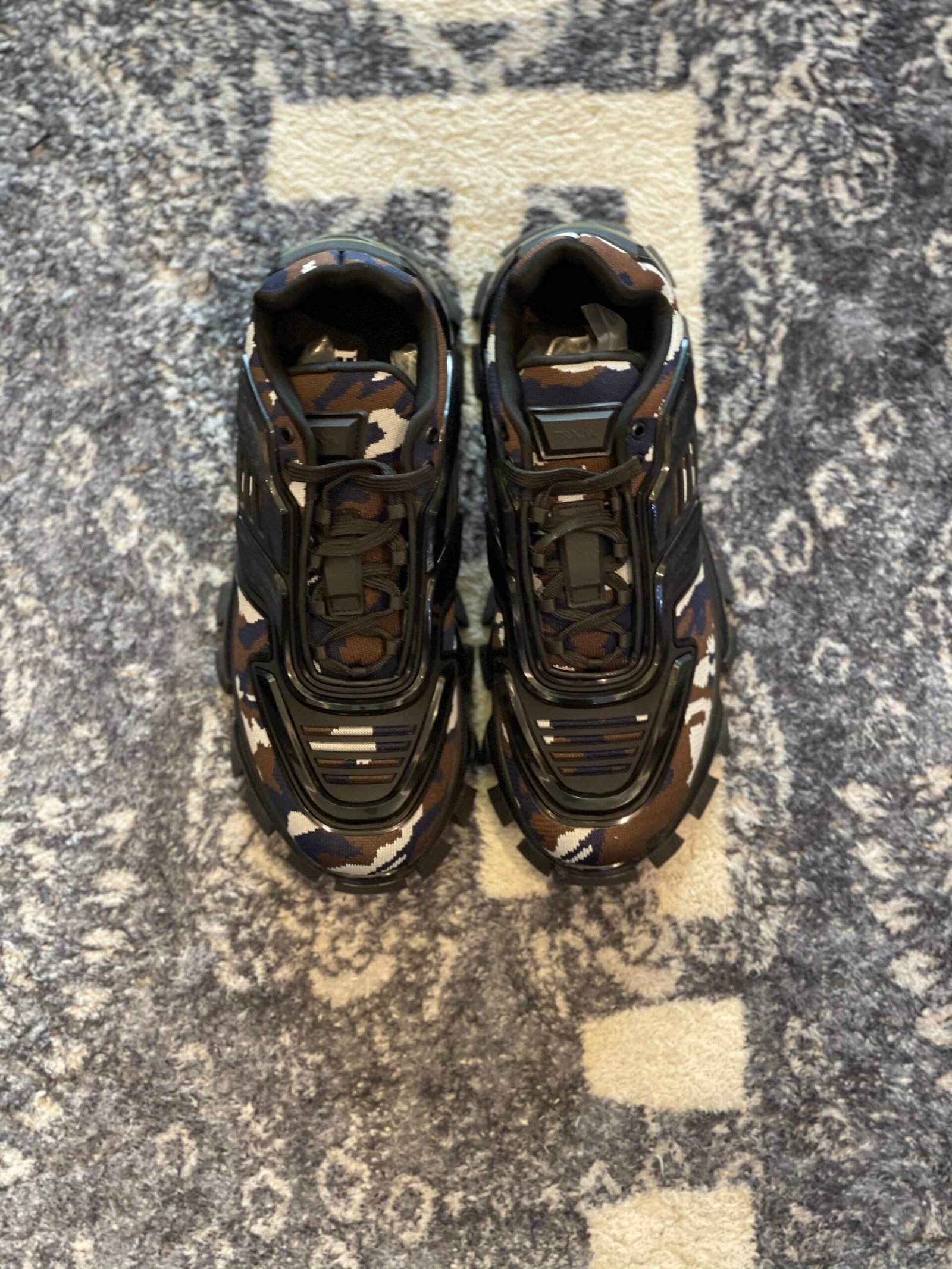 Top-down view of chunky camouflage sneakers in brown, navy, and white with black rubber industrial overlays on a patterned rug.