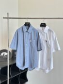 Two men’s short-sleeve button-down shirts hanging on a silver rack. One is a textured chambray blue and the other is a crisp white, both featuring a square patch logo on the front chest pocket.