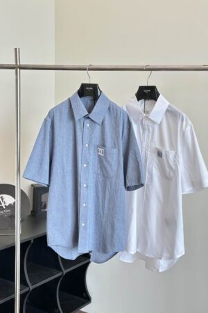 Two men’s short-sleeve button-down shirts hanging on a silver rack. One is a textured chambray blue and the other is a crisp white, both featuring a square patch logo on the front chest pocket.