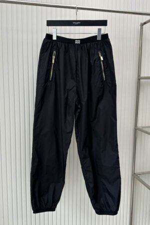 A pair of black technical track pants hanging on a rack, featuring an elastic waistband with a white Miu Miu logo, vertical gold-toned zippered pockets, and elasticized ankle cuffs.