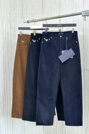 Three pairs of men's fine-wale corduroy trousers hanging on a silver rack in navy blue, black, and tobacco brown, each featuring a silver-tone enamel triangle logo on the front coin pocket.