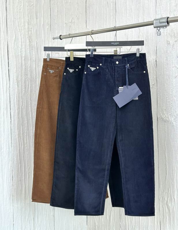 Three pairs of men's fine-wale corduroy trousers hanging on a silver rack in navy blue, black, and tobacco brown, each featuring a silver-tone enamel triangle logo on the front coin pocket.