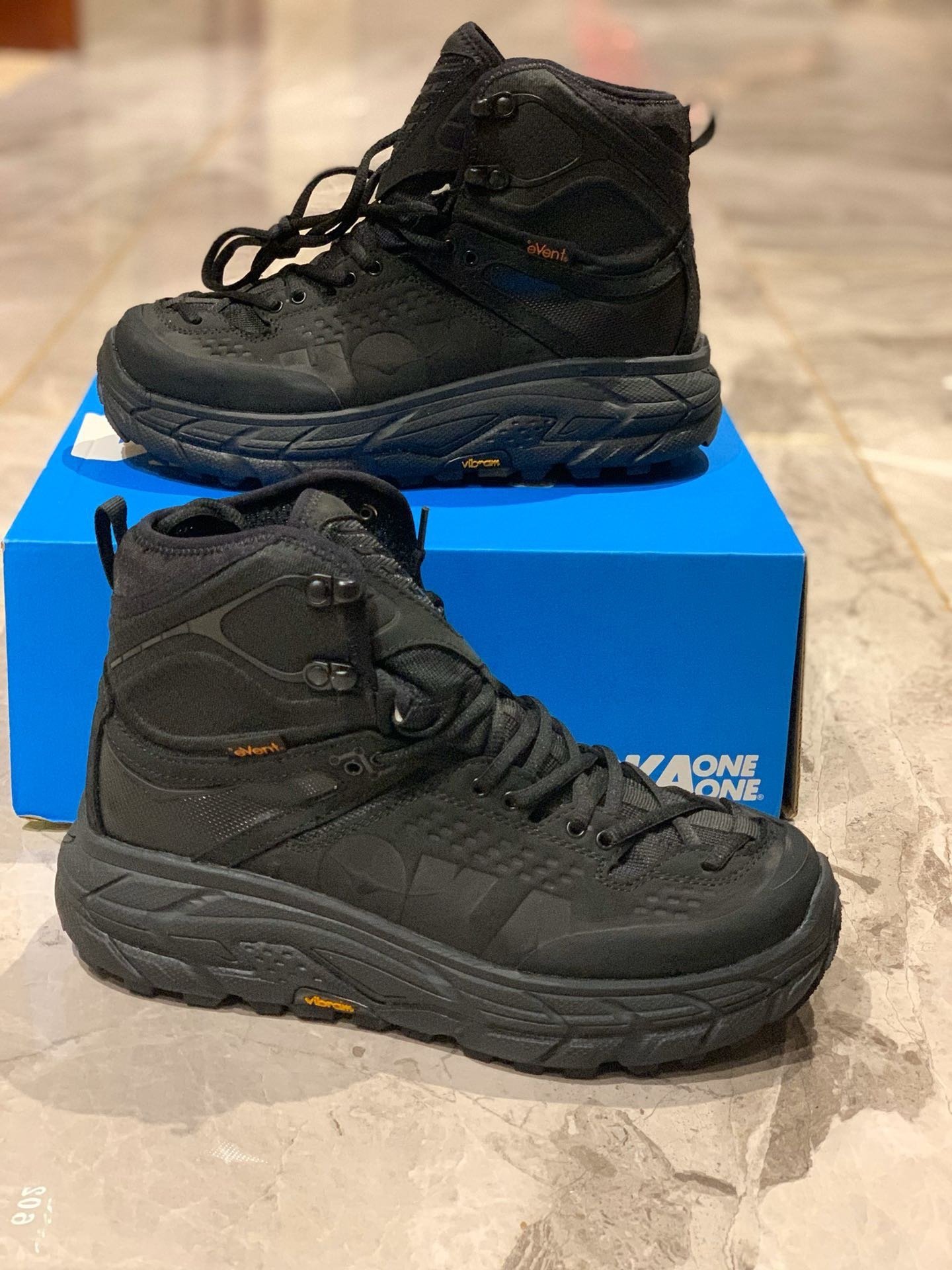 A pair of all-black Hoka One One x Engineered Garments Tor Ultra Hi boots featuring thick textured midsoles, high-top ankle support, and yellow Vibram branding, resting on a marble floor in front of a blue Hoka box.
