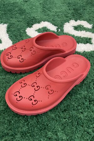 A top-down view of vibrant red rubber slip-on mules featuring perforated interlocking logos, resting on a green textured rug.