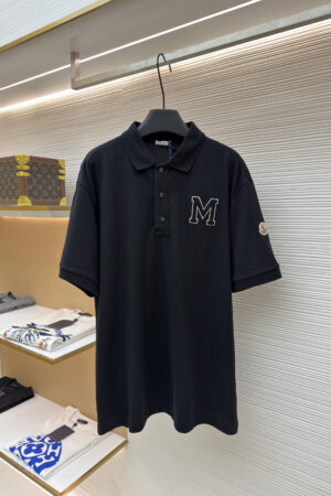 A navy blue men's pique polo shirt featuring a cream-outlined varsity "M" on the chest and a designer sleeve patch hanging on a rack.
