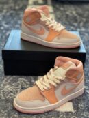 Two pairs of apricot and peach-colored Air Jordan 1 Mid sneakers displayed with one pair sitting on a black shoe box.