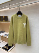 A pistachio green oversized crewneck knit sweater with a white Ami de Coeur heart logo on the left chest, hanging on a black hanger in a luxury retail environment.