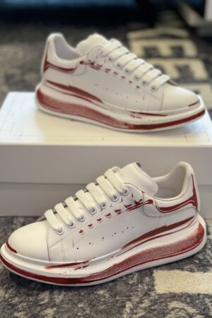 White Alexander McQueen oversized sneakers with red artistic sketch details on the sides and soles sitting on a white box.