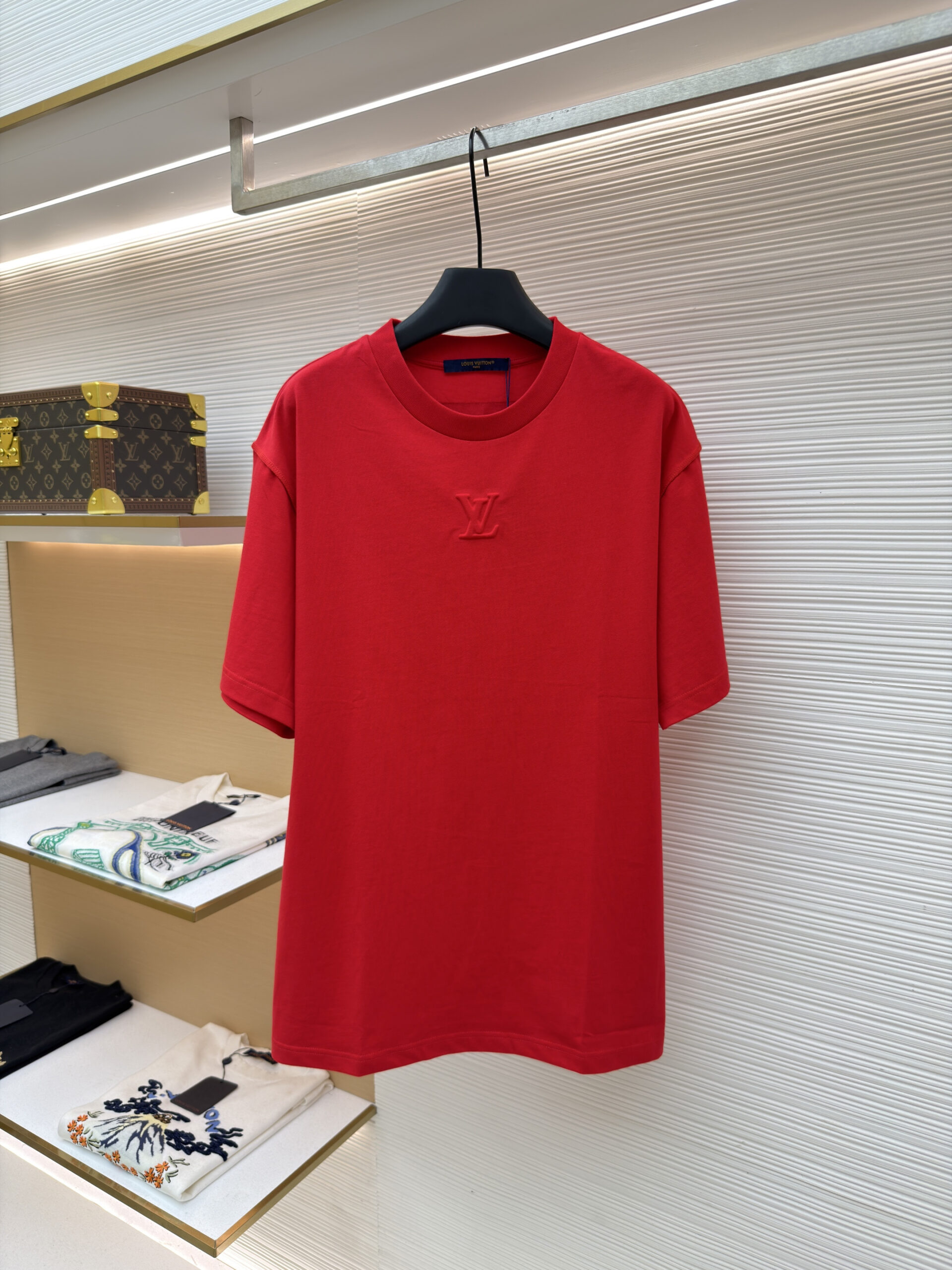 A premium men's vibrant red cotton t-shirt featuring a tonal "LV" logo embroidered on the center chest hanging on a display rack.