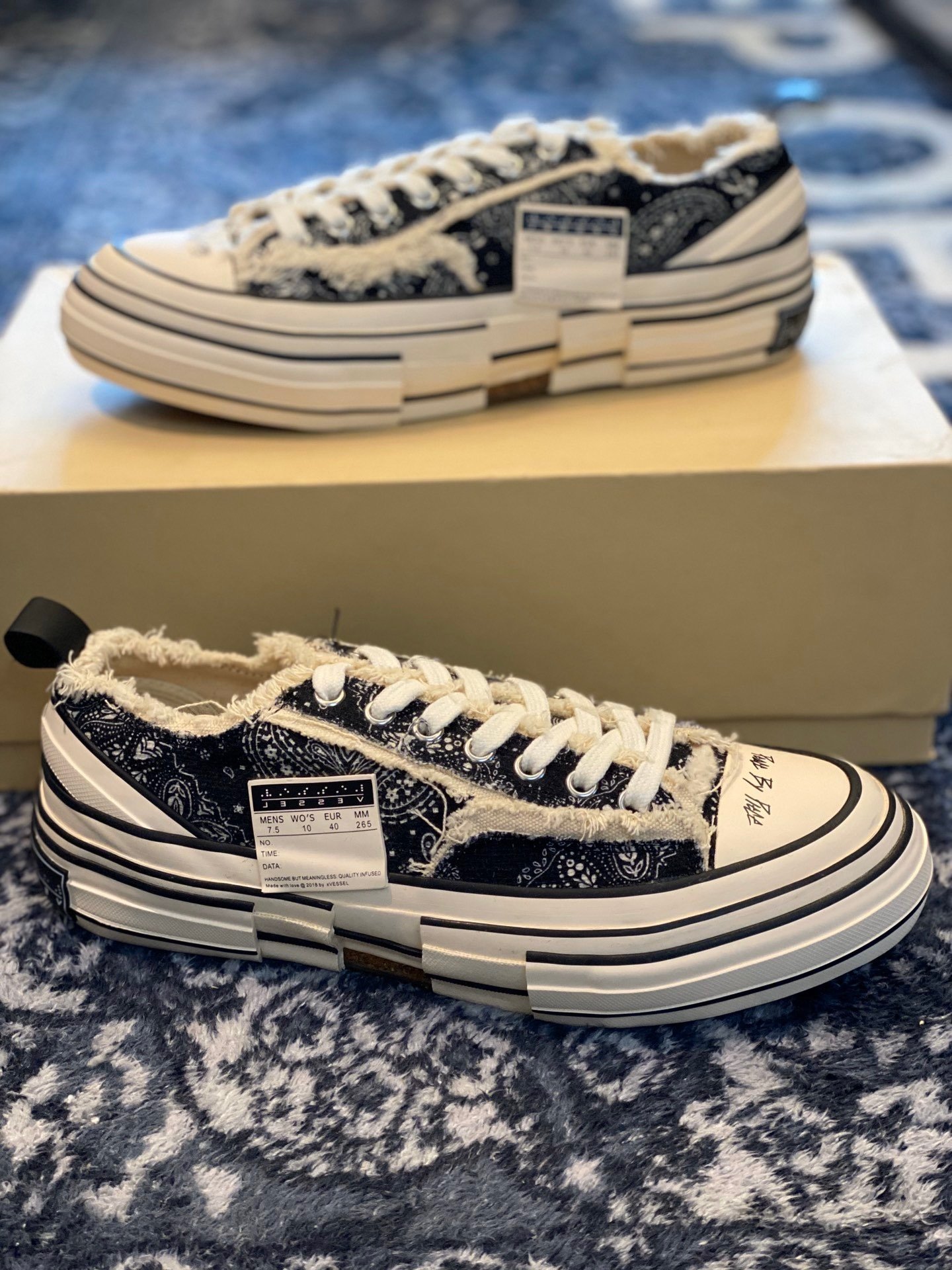 A pair of Maison Mihara Yasuhiro Peterson sneakers featuring a black and white bandana paisley print, chunky warped white midsoles, and frayed edges, resting on a blue patterned rug.